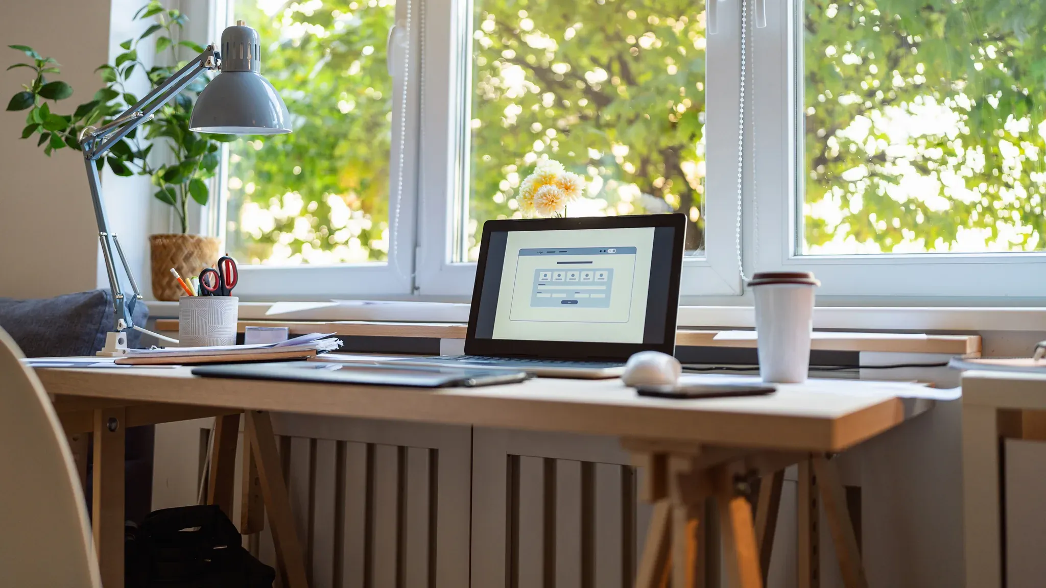 A desk with a laptop and a lamp beneath a window looking out onto trees
