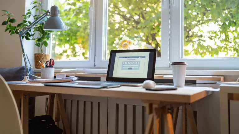 A desk with a laptop and a lamp beneath a window looking out onto trees